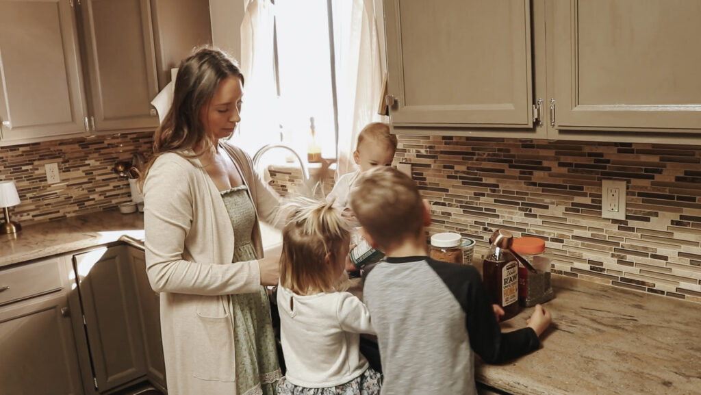Homemaker and mother stands in kitchen with her  three children while wearing a long, floral dress.