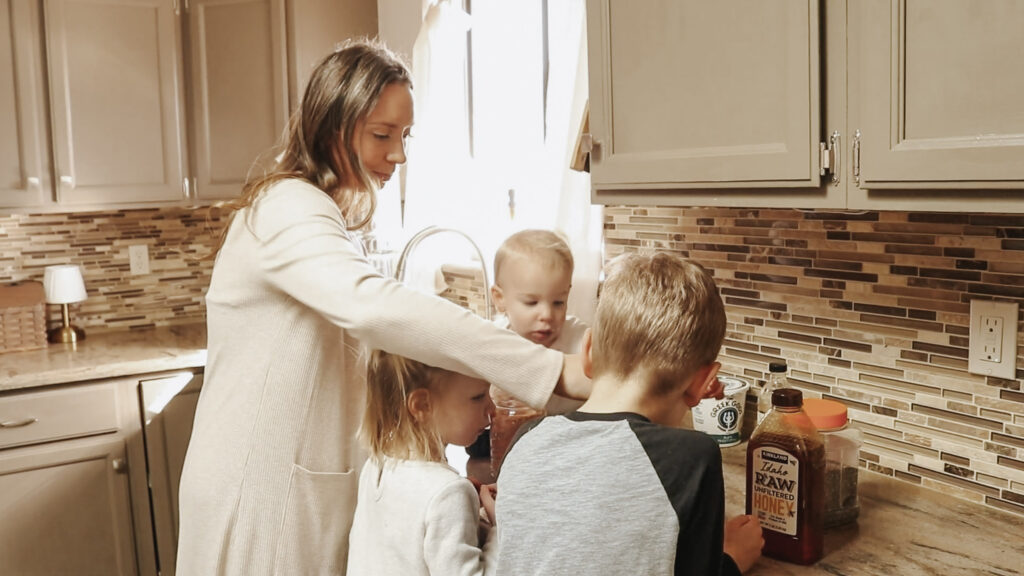 Christian homemaker and mother stands in kitchen with her children wearing a long cardigan.