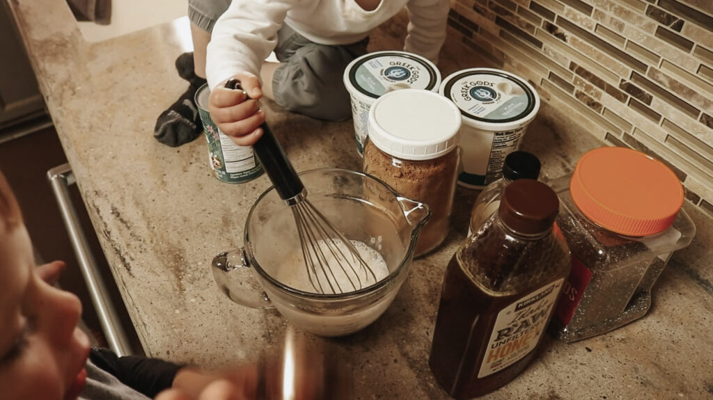 Child stirs liquid in a glass bowl with a whisk.