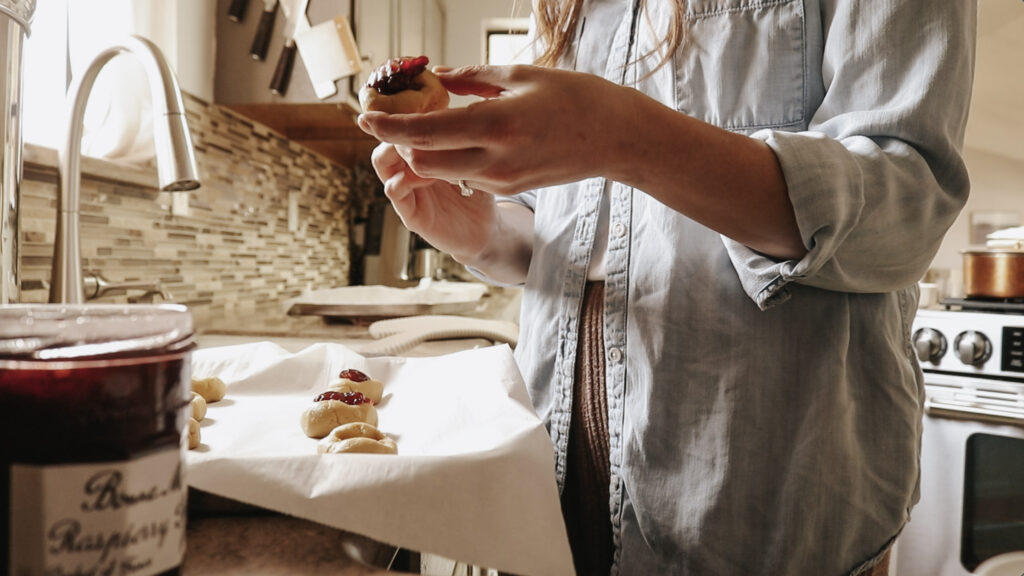 Homemaker puts jam o cookies while standing at kitchen counter.