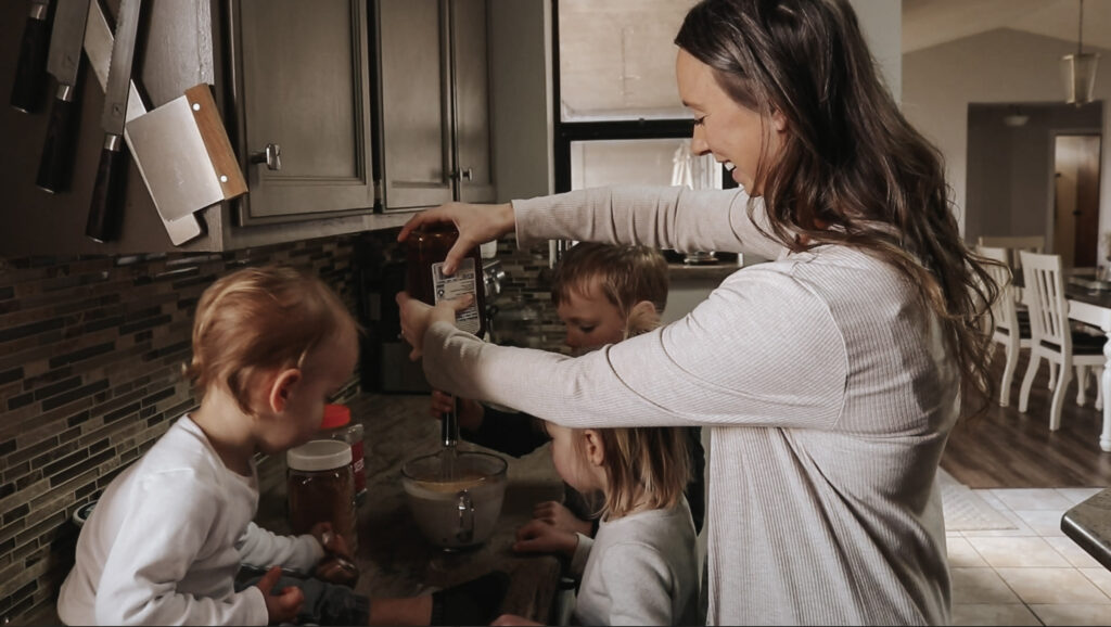 Homemaker and mother pours ingredients into bowl surrounded by her children in kitchen.