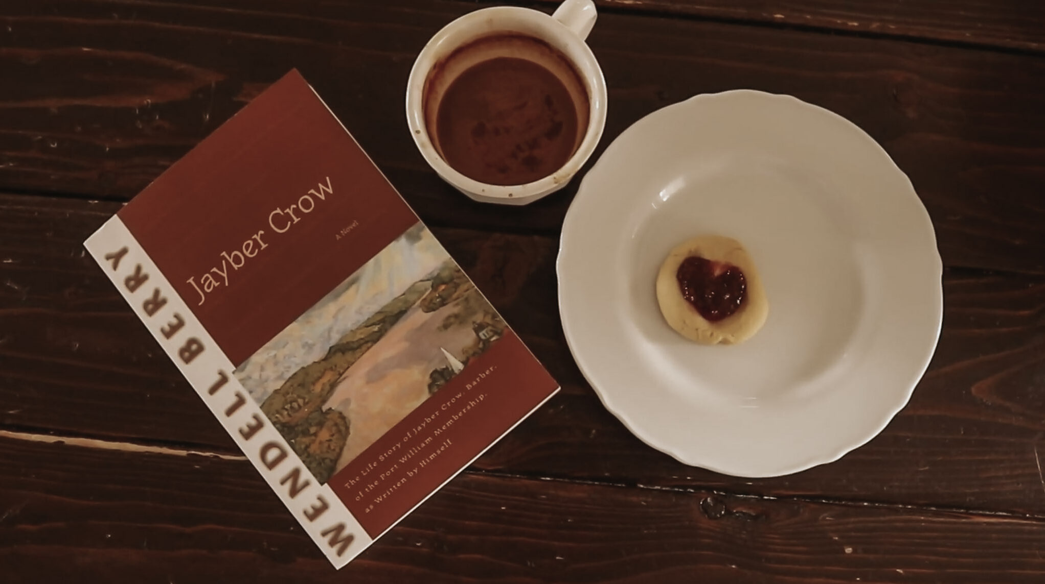 Picture of a book, cup of coffee, and small cookie on a white plate sit on a dark, wooden table.