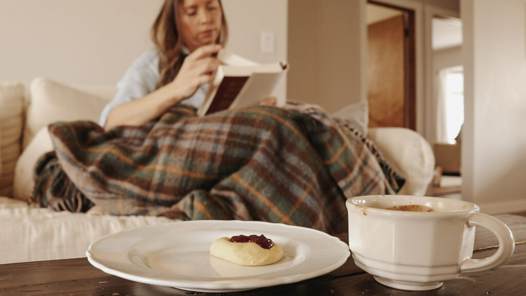 Christian homemaker sits on couch under plaid blanket while reading a book with coffee and a cookie on a coffee table.