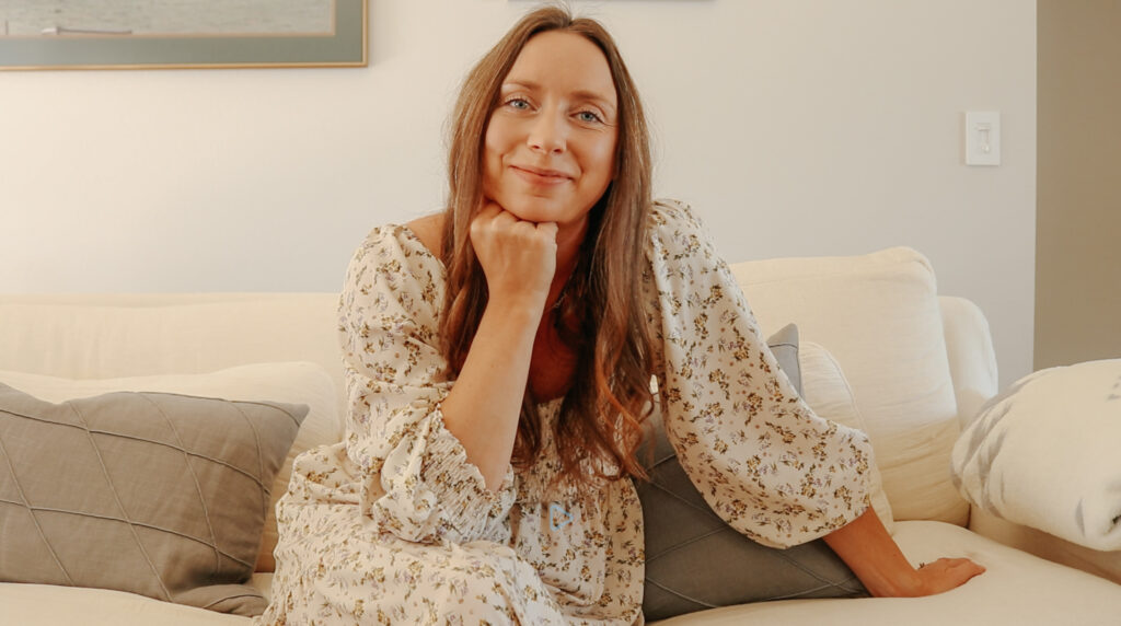 A calm and peaceful Christian homemaker in a long, smocked, floral, puff-sleeved dress props her arm on her knee and her chin in her hand. She warmly looks at the camera with a kind smile and welcoming expression.