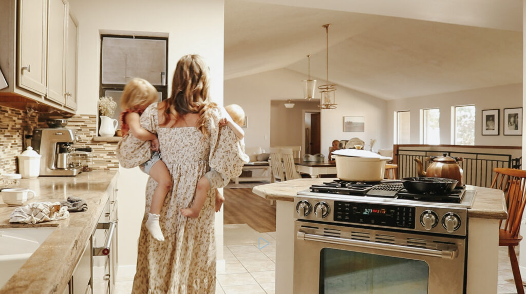 There is a Christian homemaker in a long, smocked, floral, puff-sleeved dress holding her two small children, one on each hip. She is passing through a kitchen where she has evidently been cooking and preparing a meal. There is a white enamel cost iron pot, a black cast iron pan, and a copper kettle on the stove.