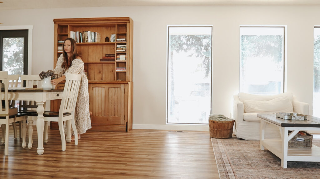 Soft morning light comes through the windows and reflects off warm wooden floors. A Christian homemaker in a long, smocked, floral, puff-sleeved dress arranges a milk white vase with purple spring flowers on a white farmhouse table. The image and atmosphere is peaceful, calm, and is evident of a morning routine completed bore 10AM.