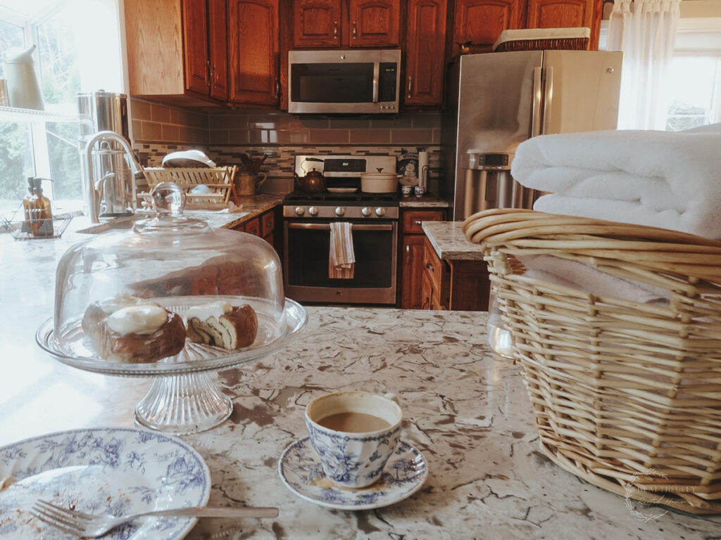 On a kitchen counter is a basket of folded blankets next to a half drunk tea cup and a plate that has evidence of a delicious dessert that has been eaten. There are cinnamon rolls iced with white cream cheese frosting in a glass pedestal display case next to the plate.
