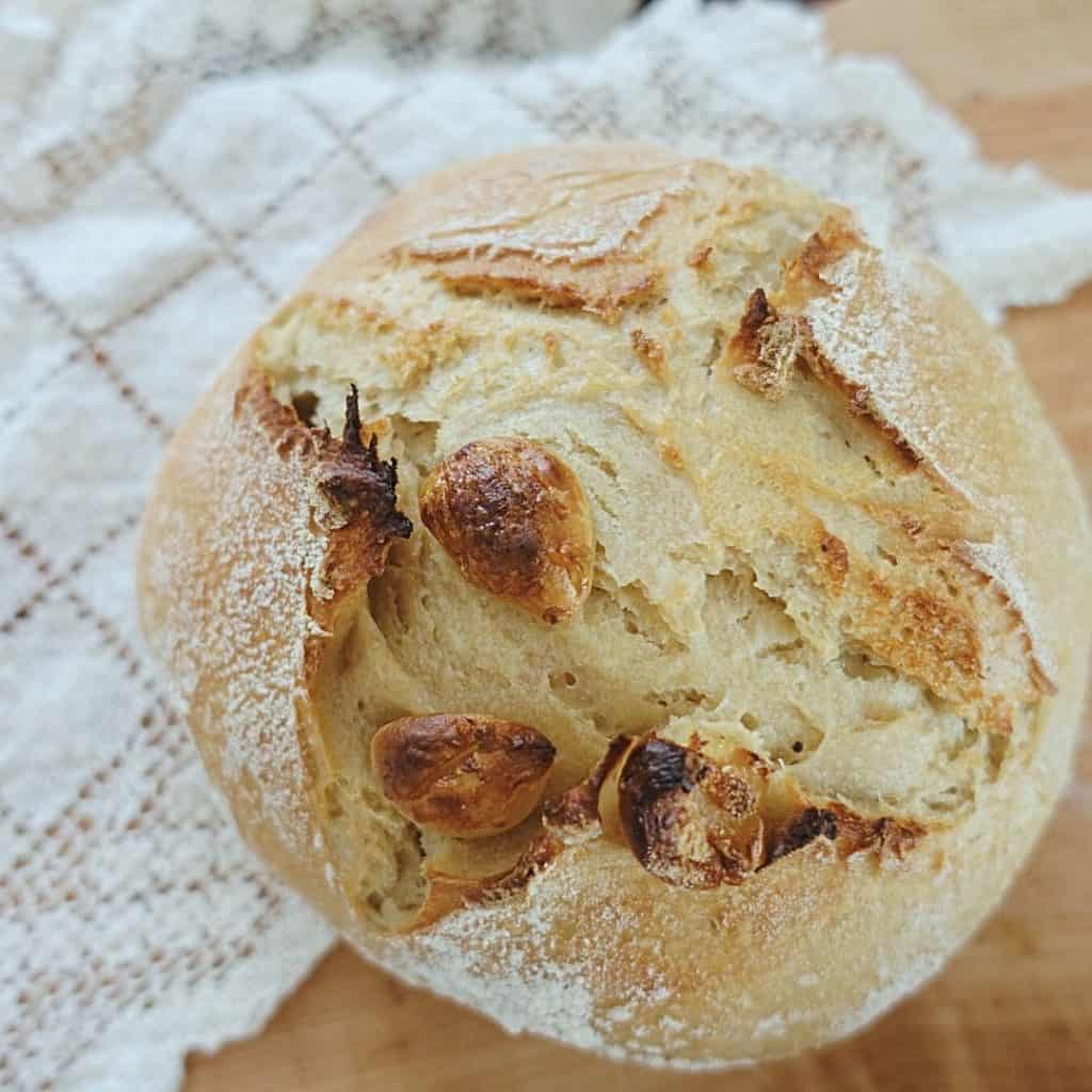 loaf of roasted garlic sourdough bread with a cross scoring pattern on top of lace doily with garlic cloves on top