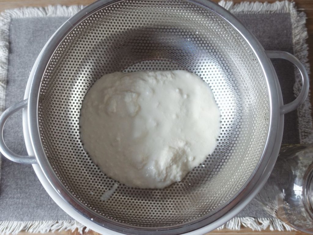 raw kefir inside of a strainer on top of a grey jute placemat with mason jar on side