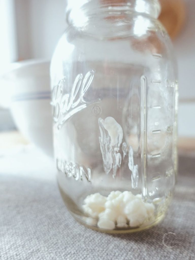 kefir grains inside a mason jar on top of a grey jute placemat in front of a ceramic bowl with blue stripes