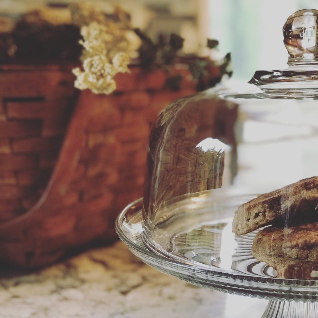 basket with stems on top and a cake stand with homemade english muffins inside on a granite countertop