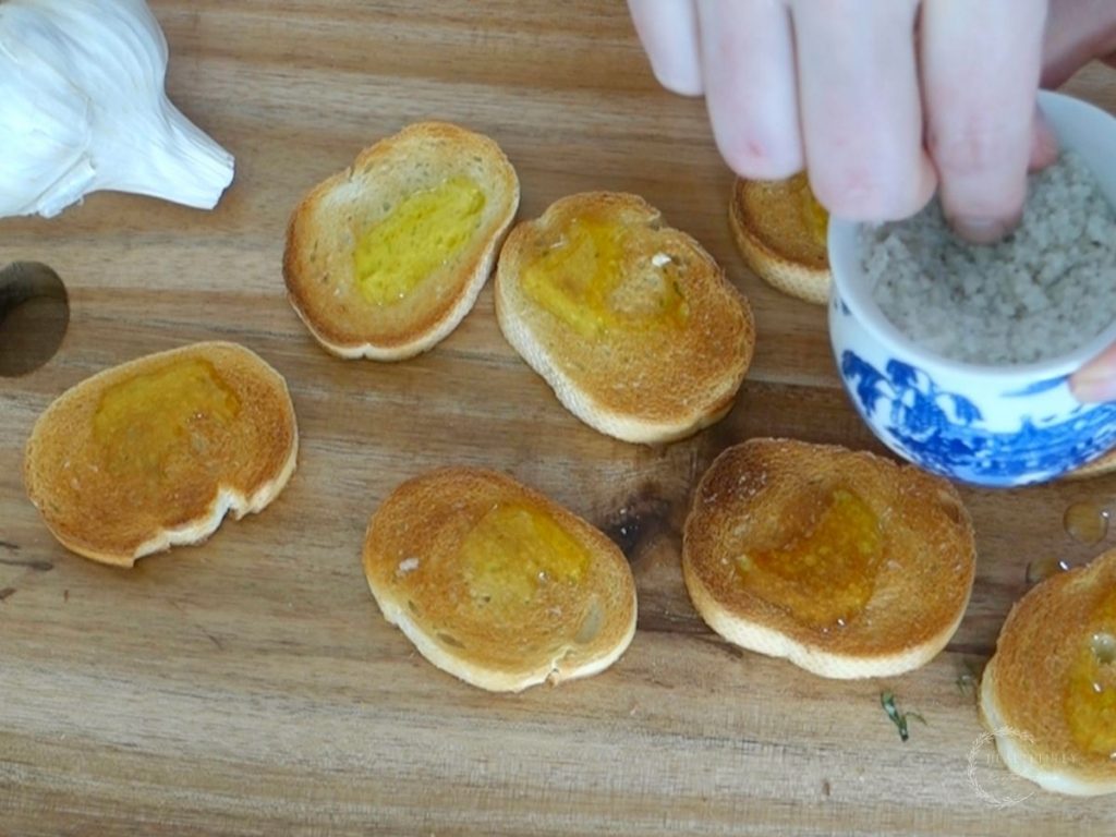 sprinkling sea salt on toasted crostini with garlic bulb on the side