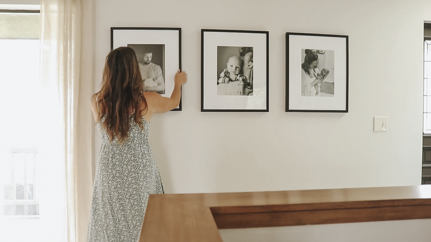 A Christian homemaker in a pale green dress with white flowers arranges a photo frame on the wall of a black and white picture of her husband hold one of her children.
