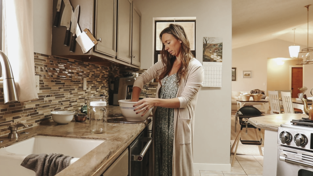 A Christian homemaker in a pale green dress with white flowers and a tan cardigan preps food in a bowl.