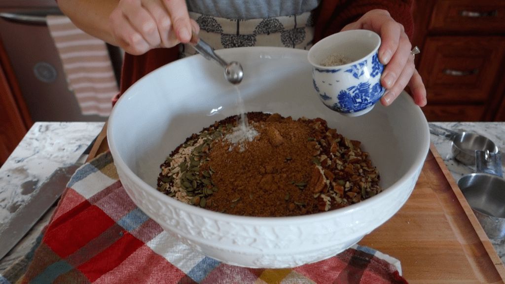 mixing the dry ingredients for pumpkin spice granola bark in a large white bowl on top of a boos block wooden butcher block sing stainless steel measuring spoons with a red checkered tea towel