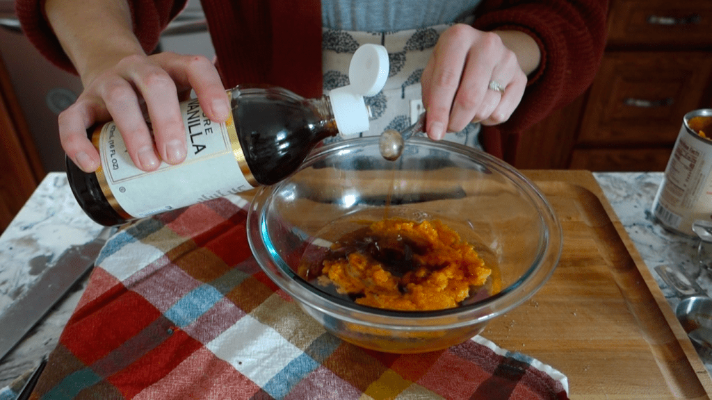 adding vanilla extract to the pumpkin puree on in a glass bowl on top of a butcher block with red checkered tea towel