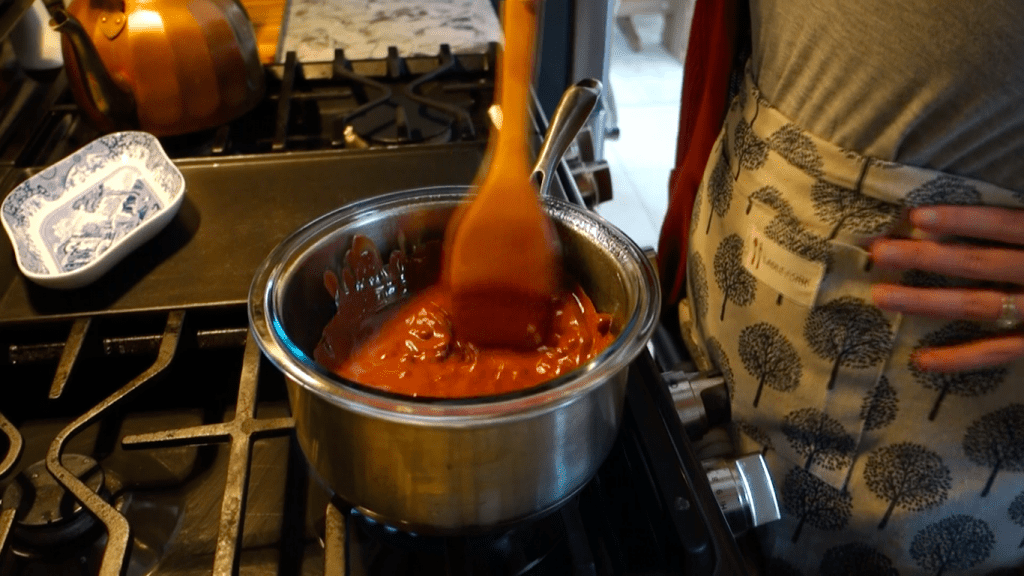using a double broiler to melt chocolate and coconut oil with a copper tea kettle and blue and white china plate on the stove wearing an apron with trees on it.