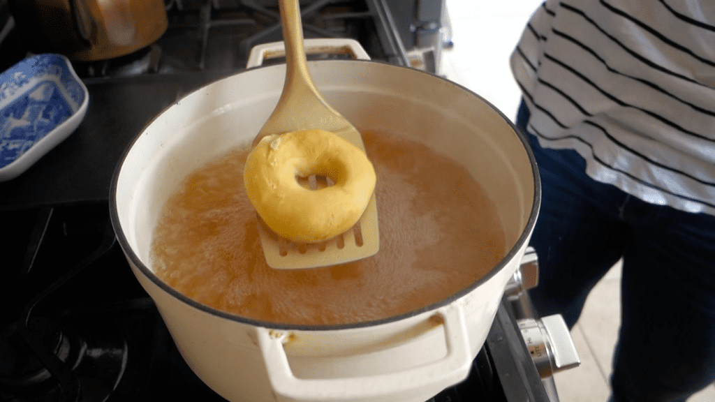 removing sourdough bagel from boiling water in a white martha stewart dutch oven on a gas stove