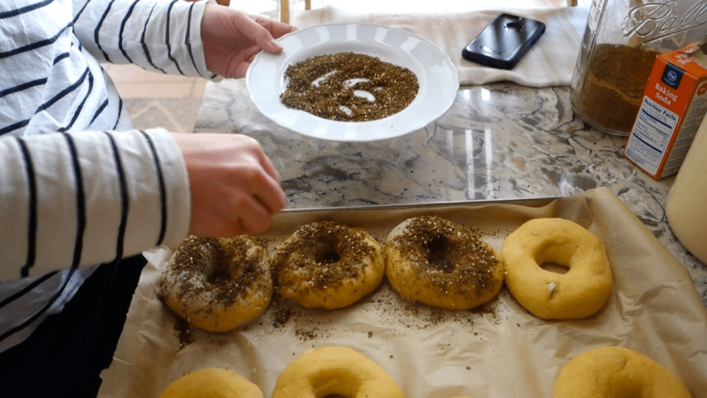 sprinkling zaatar seasoning onto homemade sourdough bagels which are sitting on a parchment lined baking sheet