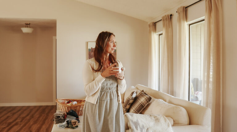 A Christian homemaker in a green and white checkered dress holds a white coffee cup and gazes out the window.