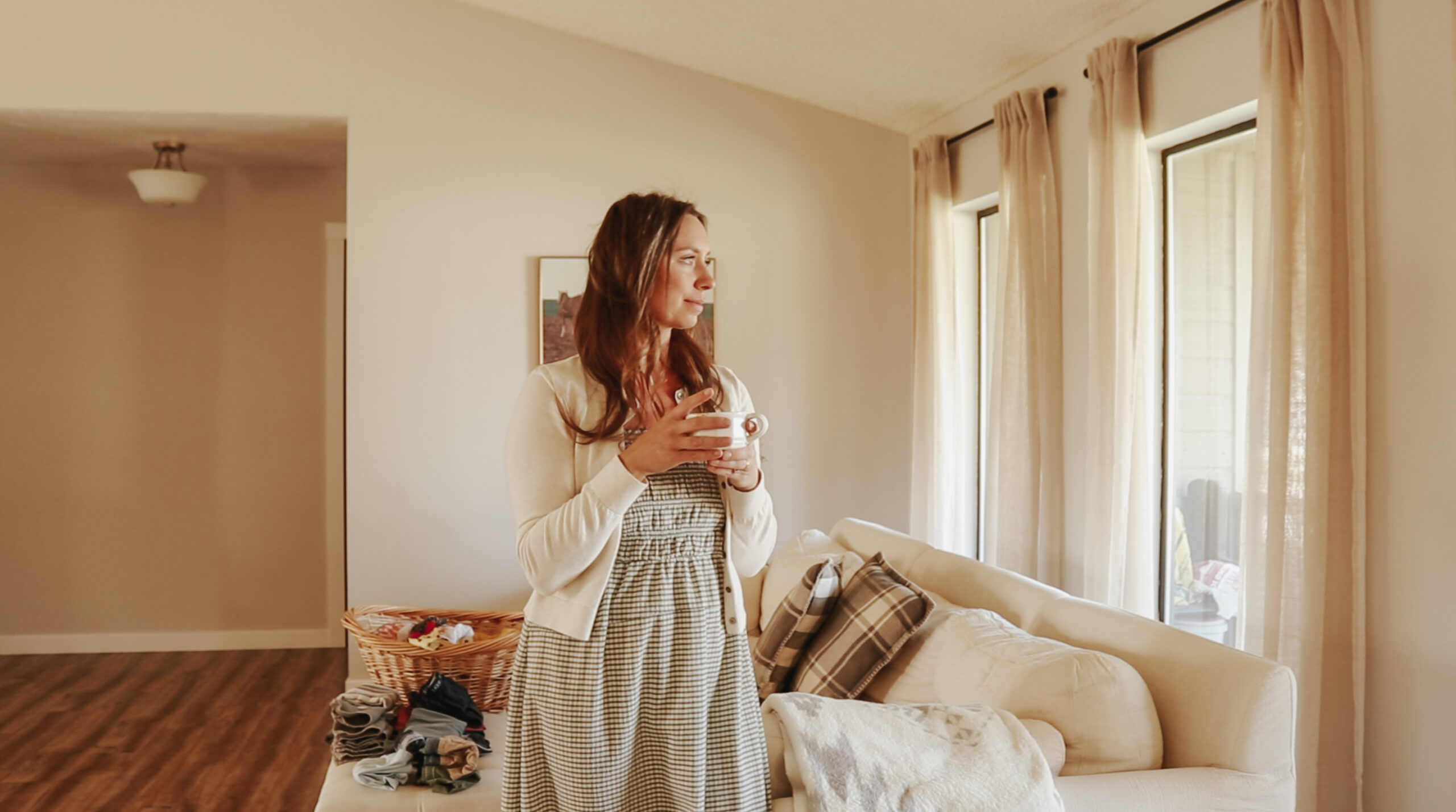 A Christian homemaker in a green and white checkered dress holds a white coffee cup and gazes out the window.