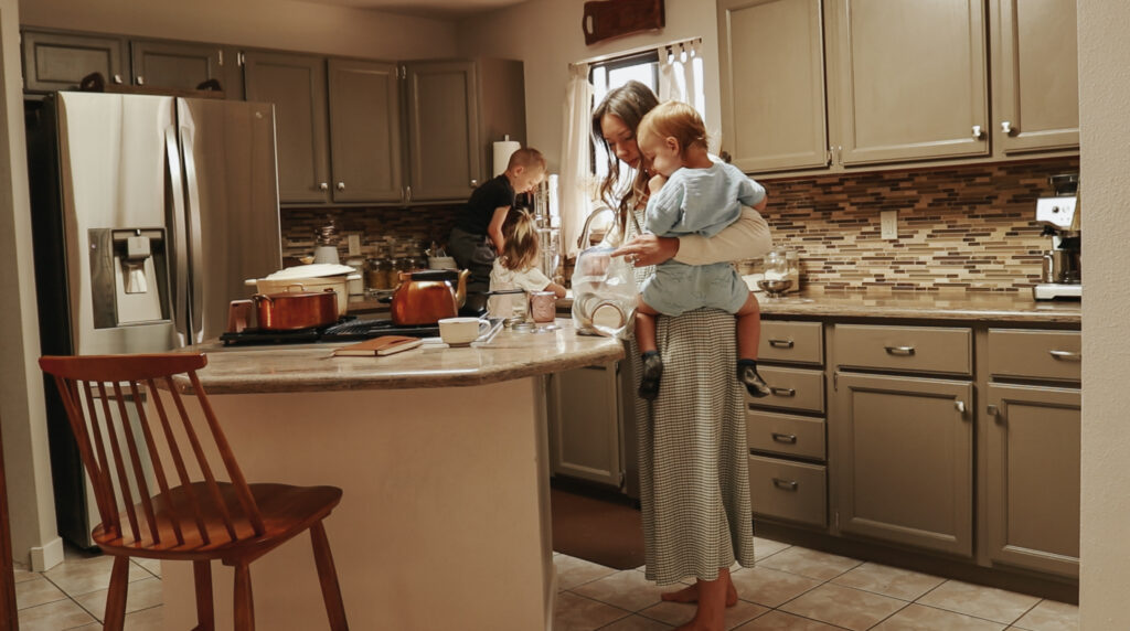 A Christian homemaker in a green and white plaid dress and white cardigan looks through a plastic bag while she holds her youngest son on her hip.