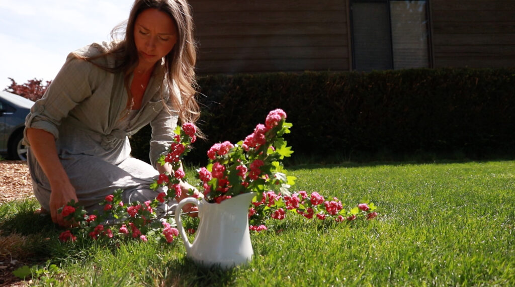 A Christian homemaker in a gray dress and green shirt kneels on the grass as she arranges a bouquet of pink flower in a white vase.