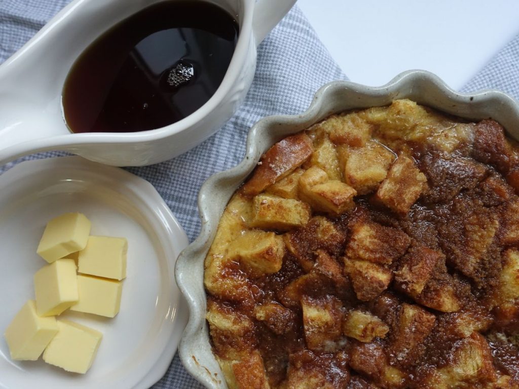 close up of brioche french toast casserole after being baked inside of a scalloped baking dish with a side of syrup and butter