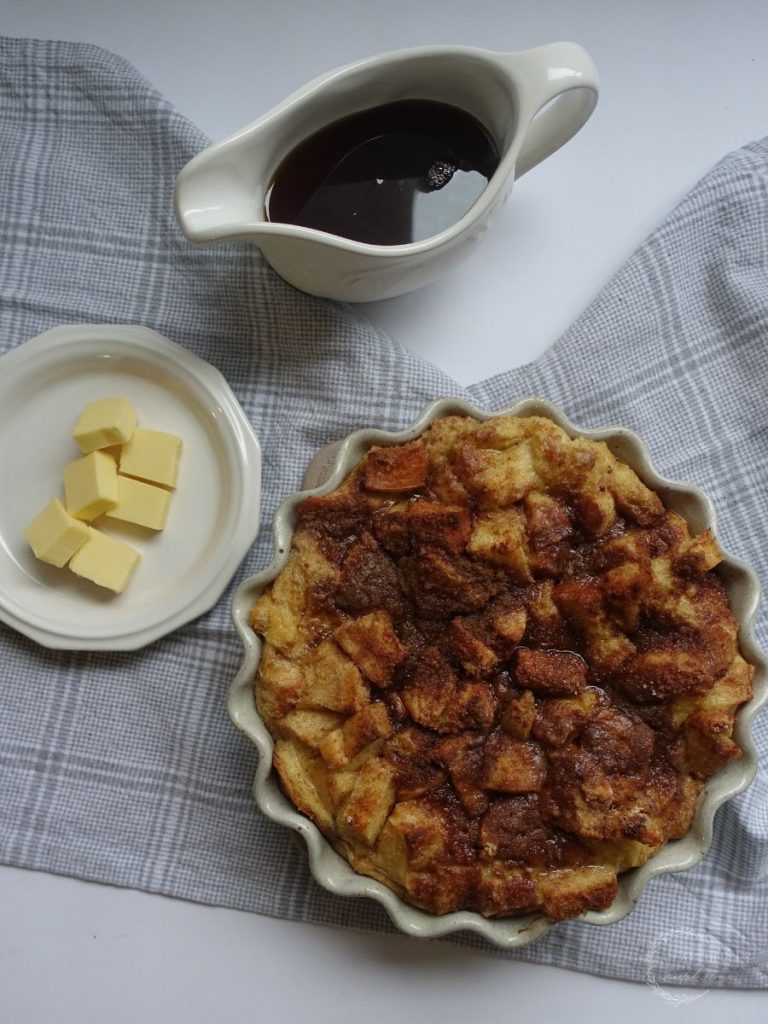brioche french toast casserole in a round scalloped dish with a plate of butter pads and jar of maple syrup next to it