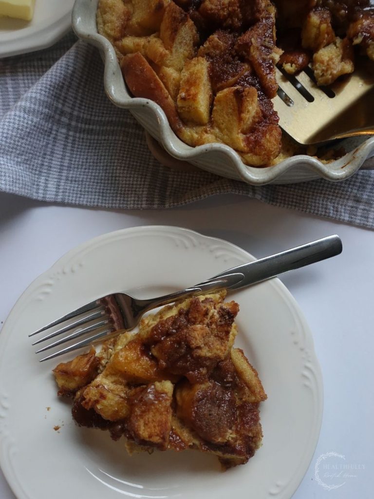 slice of brioche french toast casserole on a plate with a fork and the casserole dish next to it with a gold spatula coming out of it