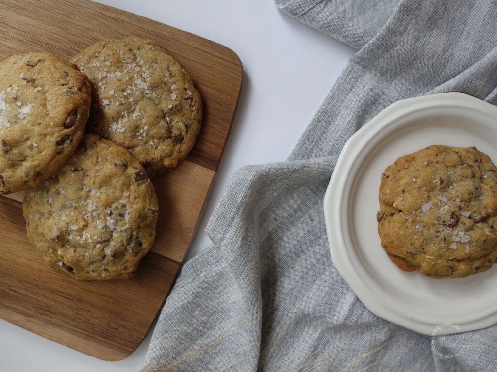 close up of chocolate toffee cookies without brown sugar on a wooden serving platter with another one on a white plate showing how large they are