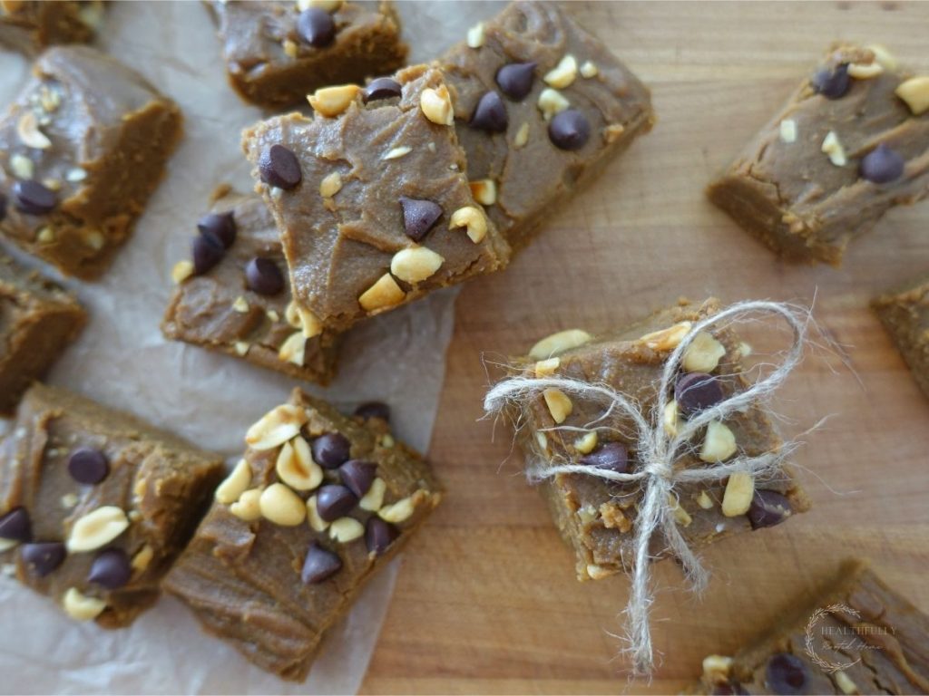 peanut butter fudge pieces topped with peanuts and chocolate chips scattered on a wooden cutting board with a couple wrapped in twine