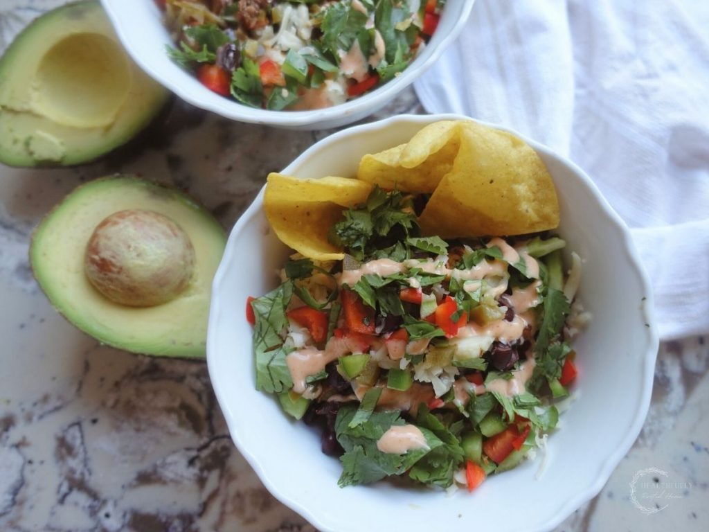 deconstructed taco in a white bowl with tortilla chips inside and a halved avocado on the side