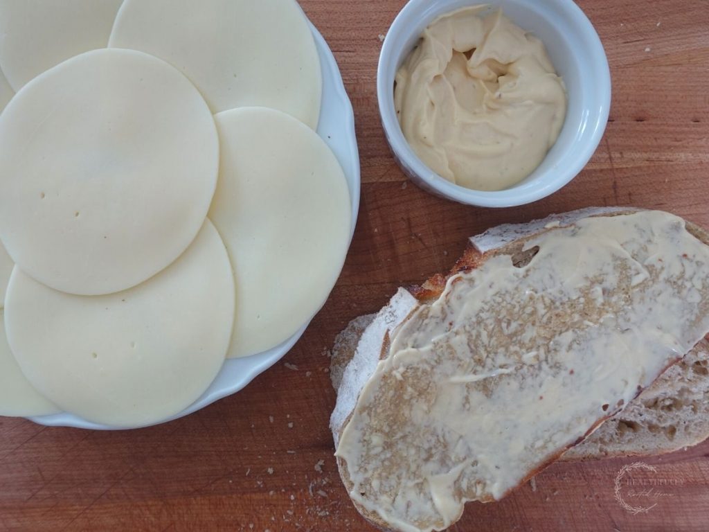 slice of sourdough bread with mayo spread overtop and a plate of sliced cheese ready to make a mayo grilled cheese sandwich