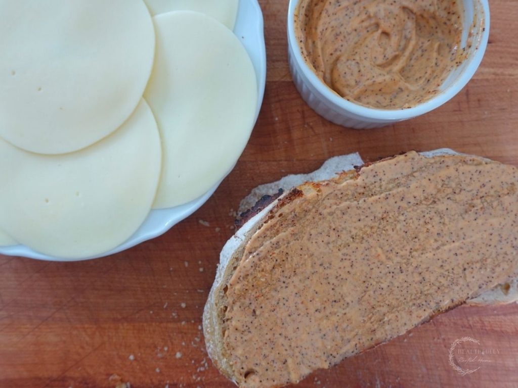 sourdough bread topped with ancho aioli with a plate of sliced cheese on the side getting ready to make ancho aioli grilled cheese sandwiches