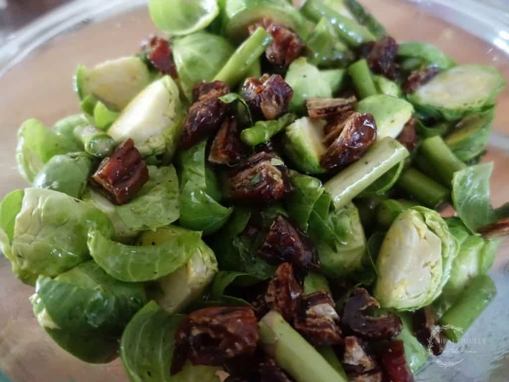 chopped asparagus and brussels sprouts with diced dates tossed with avocado oil and salt and pepper in a large glass bowl