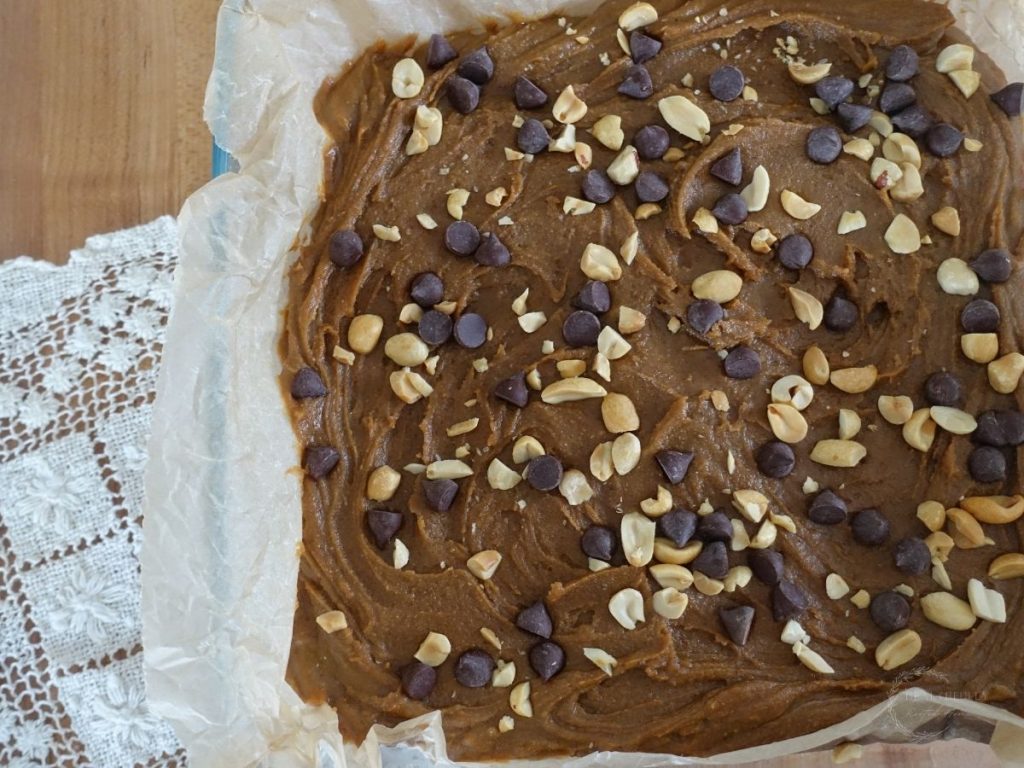 peanut butter fudge in a baking dish topped with chopped peanuts and chocolate chips next to a lace doily
