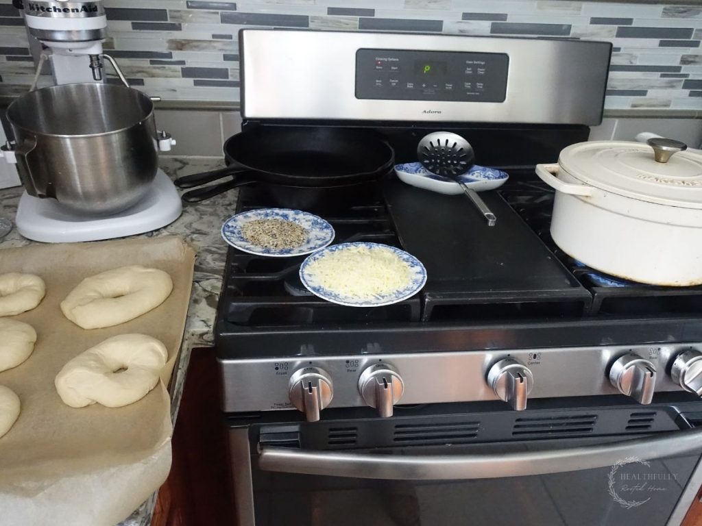 kitchen prepped for making sourdough discard bagels with a white dutch oven of boiling water and shaped bagels on a parchment lined baking sheet and toppings on the side