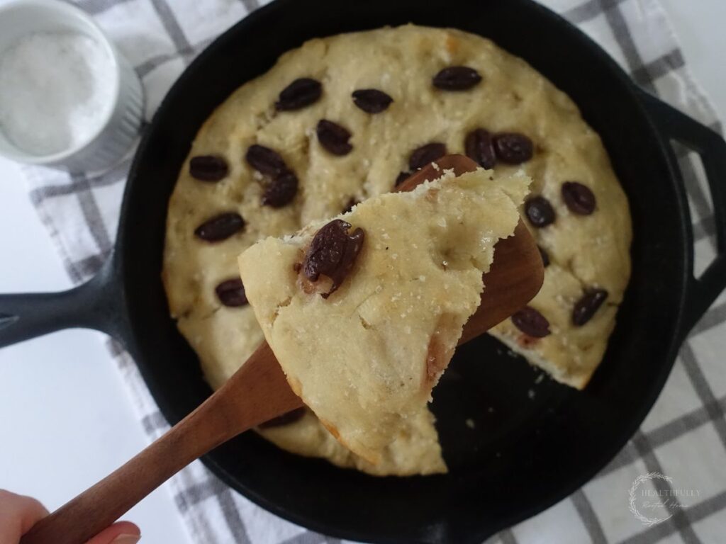 slice of sourdough discard focaccia bread on a wooden spoon being held up overtop the entire loaf in a cast iron skillet