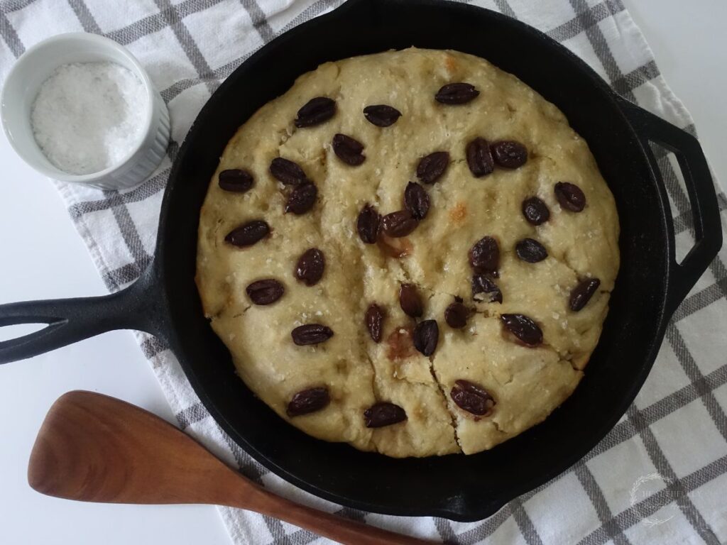SOURDOUGH DISCARD FOCACCIA bread in a cast iron skillet with kalamata olives and a wooden spoon on the side