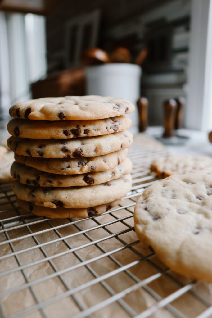 chocolate chip sugar cookies stacked on top of eachother with more on the cooling rack beside it showing the thinness of them