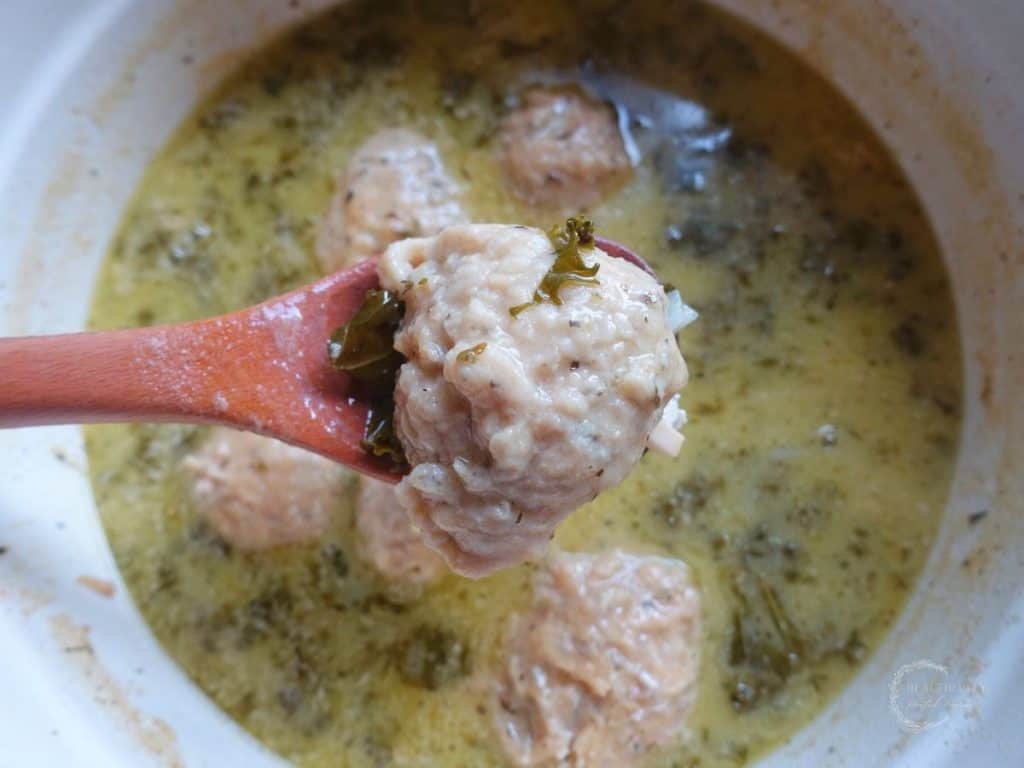 sourdough dumpling with a piece of kale on top resting on a wooden spoon over a white dutch oven full of kale chicken soup