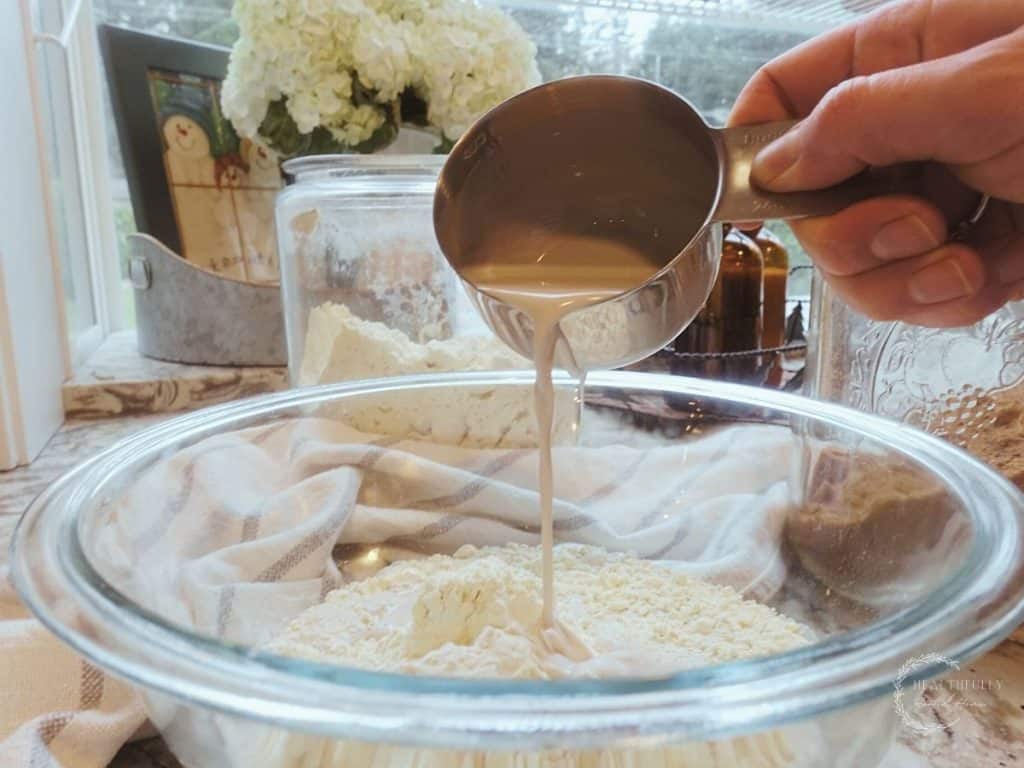 making sourdough pancake batter in a large glass bowl pouring buttermilk on top of einkorn flour with hydrangeas in background