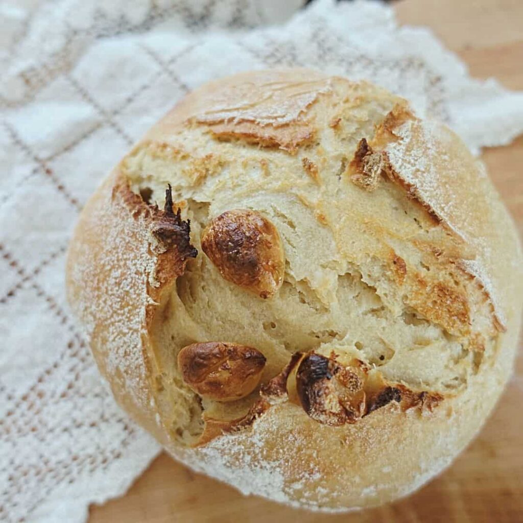 loaf of roasted garlic sourdough bread with a cross scoring pattern on top of lace doily with garlic cloves on top