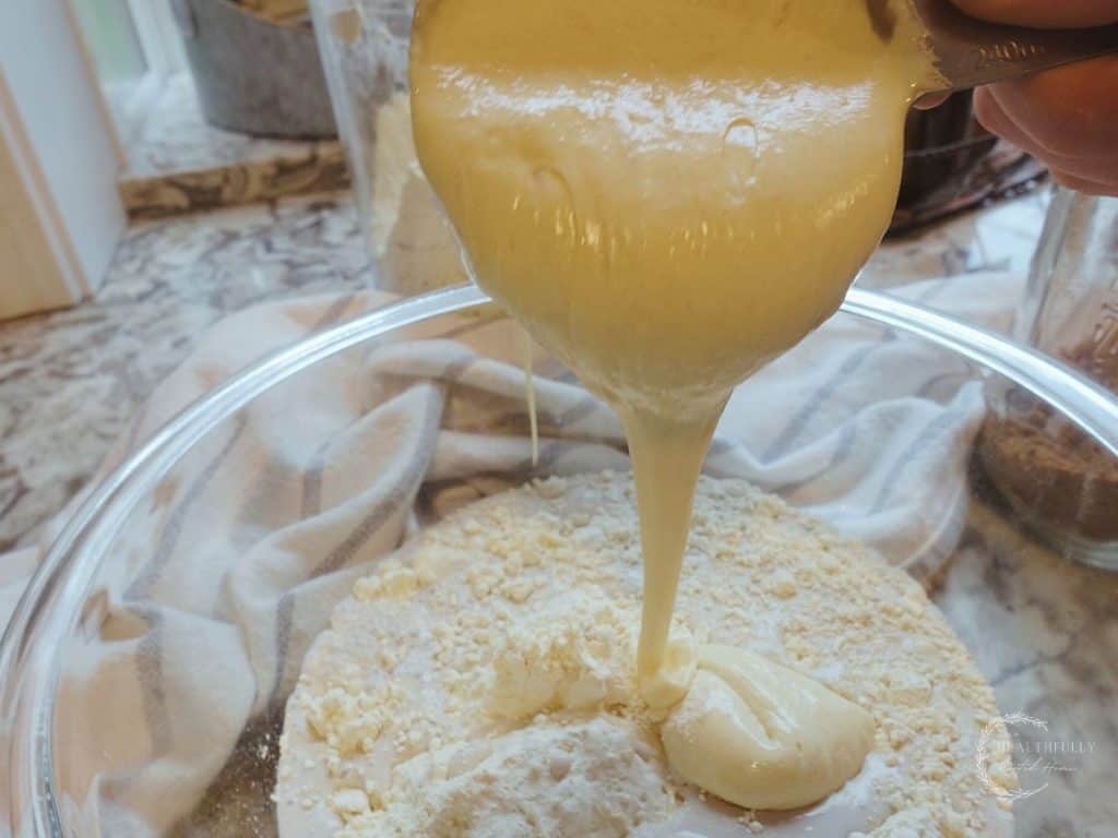 pouring sourdough starter into the pancake batter in a large glass bowl with a blue and white striped tea towel in back