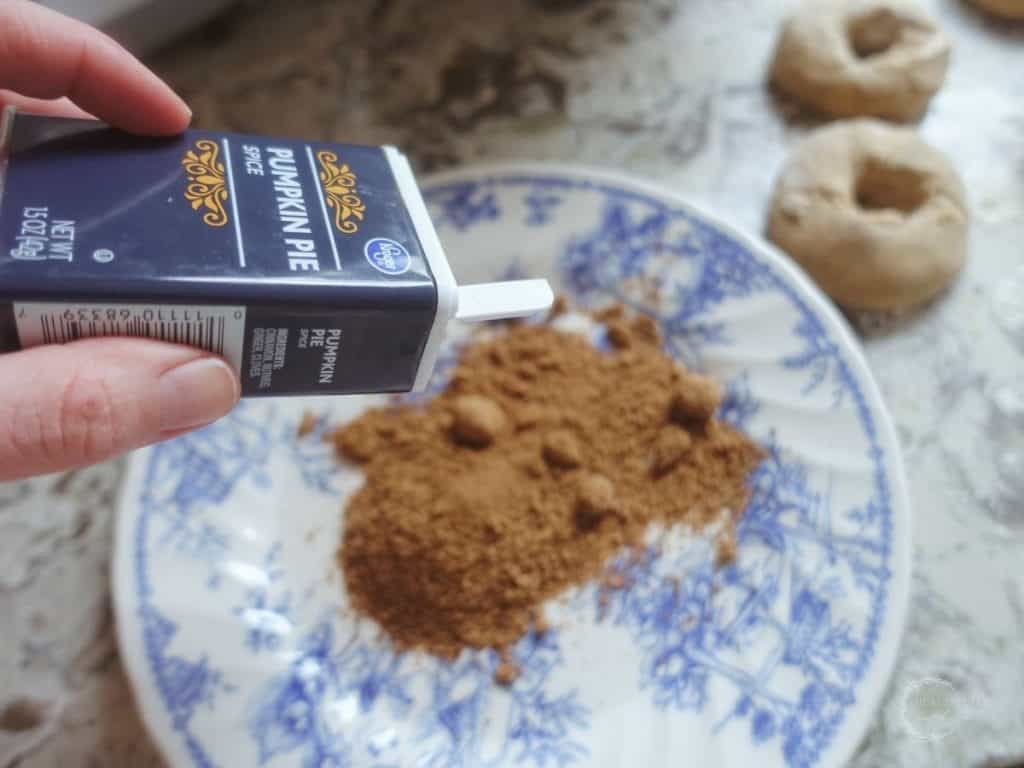 pouring pumpkin pie spice on top of coconut sugar on a blue and white plate with shaped donuts in the background