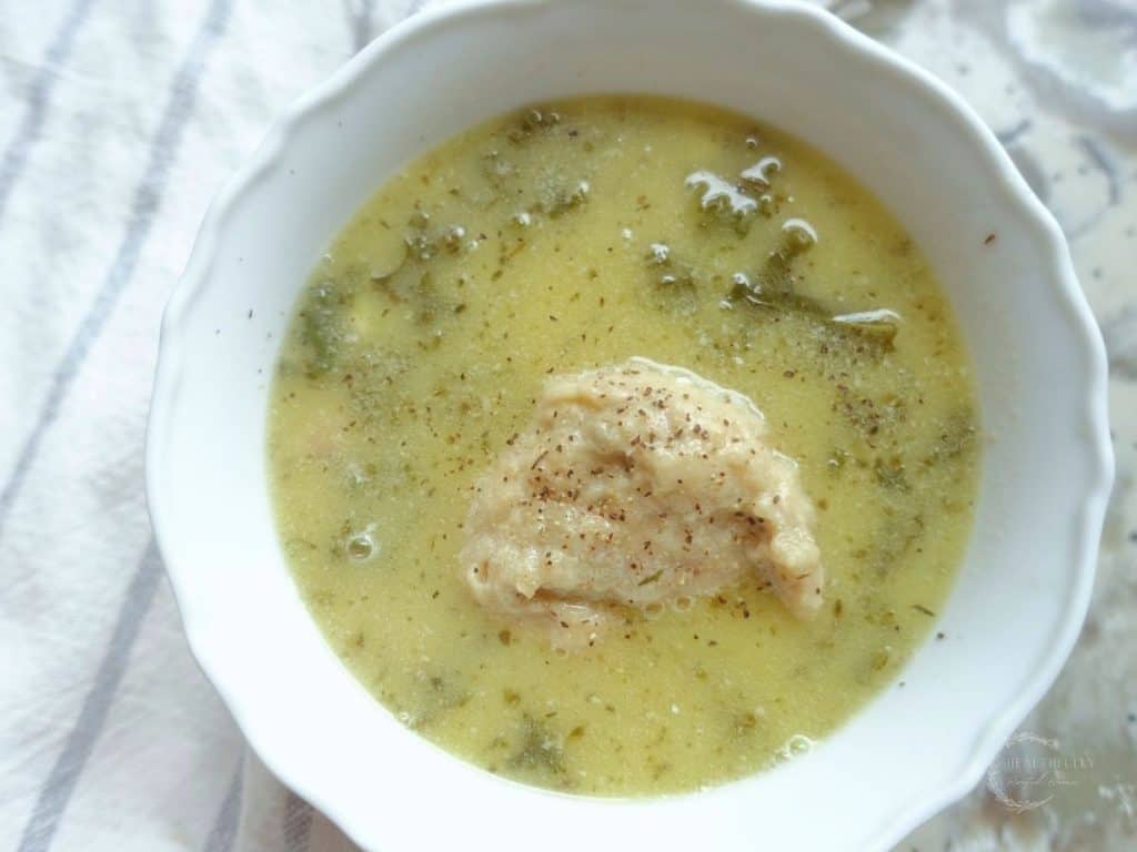 kale chicken soup with a sourdough dumpling in a white bowl and a blue and white striped tea towel on the side