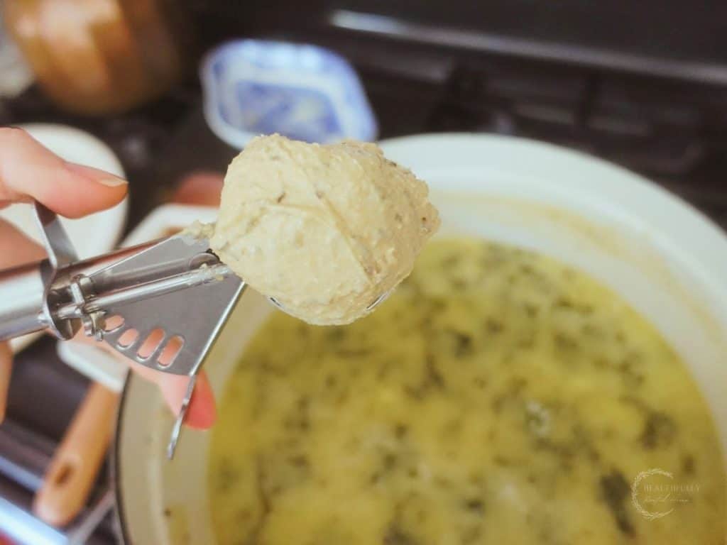 sourdough dumpling batter scooped with a cookie scoop with a dutch oven full of kale and chicken soup in the background and a blue and white china plate with copper tea kettle