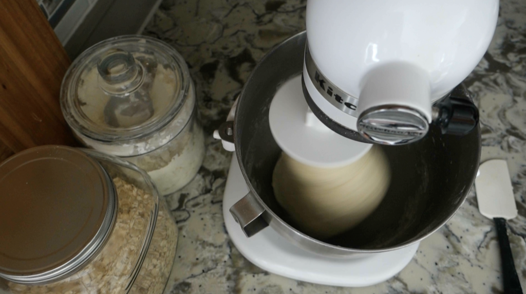white kitchenaid mixer kneading sourdough pretzel dough with glass jars of flour and oats next to it