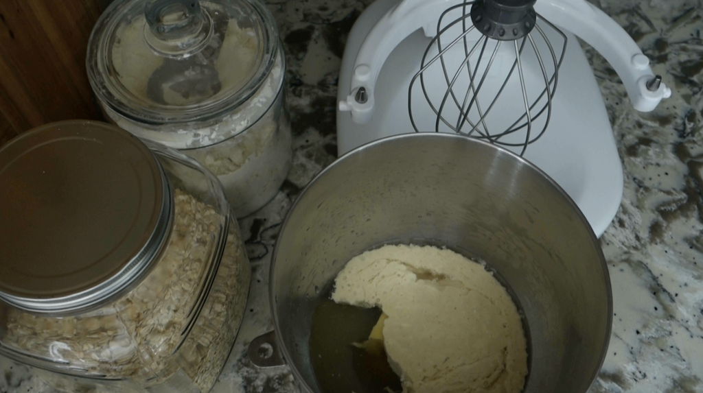 bowl of a stong mixer with the ingredients for sourdough pretzel dough next to a white kitchenaid mixer and jars of flour and oats